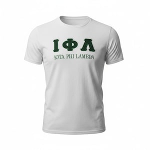 May include: White t-shirt with the Greek letters Iota, Phi, and Lambda in dark green, above the text "IOTA PHI LAMBDA". The shirt has a crew neck and short sleeves. The design is centered on the front of the shirt.