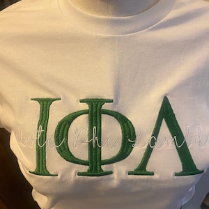 May include: White t-shirt with green embroidered Greek letters and the words "Iota Phi Lambda" in a cursive font. The letters are large and bold, with a textured appearance. The shirt is made of a soft, smooth fabric.