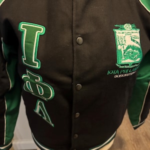 以下が含まれることがあります： 白のトリムが付いた黒と緑のレターマンジャケット。ジャケットには、白いアウトラインの緑色のギリシャ文字I、D、Aが使用されています。右胸には緑と白の紋章があり、下に「Iota Phi Lambda Sorority」の文字がプリントされています。