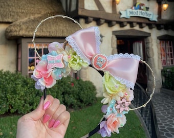 May include: Pink and white Minnie Mouse ears with a pink bow, white lace trim, and a floral arrangement. The ears have a "Eat Me" cookie charm and a "Drink Me" cookie charm.