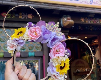 Peut inclure: Un serre-tête Mickey Mouse doré avec une couronne florale composée de fleurs violettes, roses, jaunes et blanches. Une couronne dorée scintillante est placée au sommet de la couronne florale.