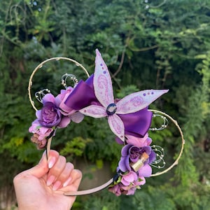 以下が含まれることがあります： 大きな紫色の蝶、紫色のリボン、紫色の花の房が特徴の装飾的なヘアバンド。 ヘアバンドには金色のワイヤーアクセントがあり、緑色の背景に対して手で持たれています。