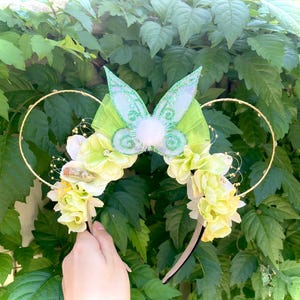 May include: A whimsical headband featuring gold wire ears, adorned with light green and white flowers, and a central butterfly design with glitter accents. The headband is set against a backdrop of lush green foliage.