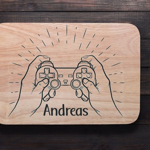 May include: A wooden cutting board with a black and white illustration of a video game controller and the name "Andreas" printed on it.