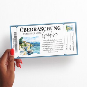 以下が含まれることがあります： 「&Uuml;BERRASCHUNG Wir machen Urlaub am Gardasee」の文字と湖の風景の水彩イラストが描かれた水色のギフト券。赤いマニキュアを塗った手でギフト券が持たれています。