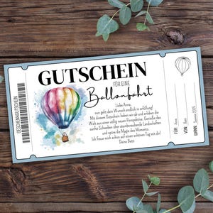 Puede incluir: Un vale de regalo azul y blanco para un paseo en globo aerostático. El vale de regalo presenta un globo aerostático colorido sobre un fondo de acuarela. El texto en el vale de regalo dice "Gutschein für eine Ballonfahrt".