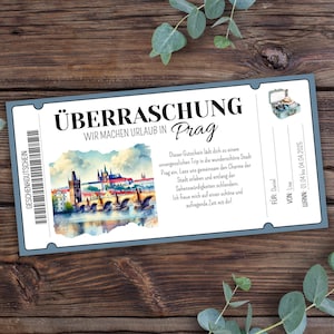 Pode incluir: Um vale-presente com o texto "ÜBERRASCHUNG Wir machen Urlaub in Prag" e uma imagem em aquarela de uma ponte e edifícios. O vale tem uma borda azul e está sobre uma superfície de madeira.