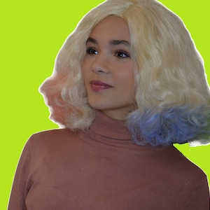 May include: A woman with blonde hair with pink and blue highlights is wearing a brown turtleneck sweater.