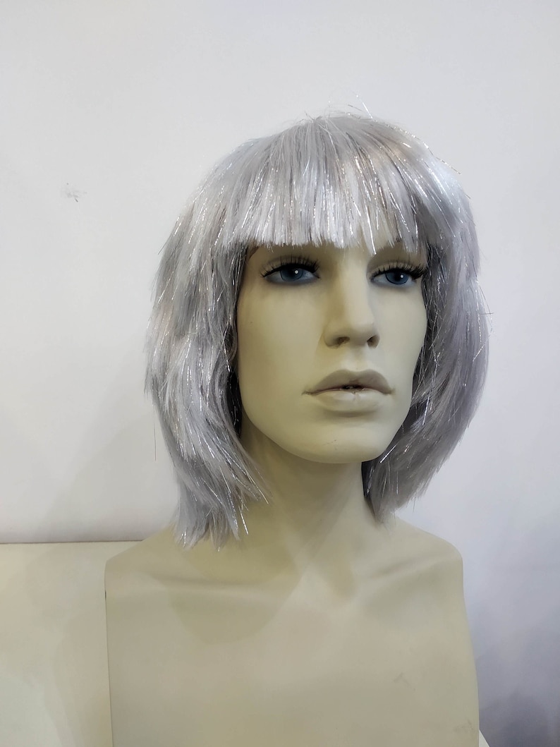 May include: A mannequin head wearing a silver wig with a blunt fringe. The wig has a short, layered style with strands of silver tinsel throughout. The mannequin has blue eyes and a neutral expression, set against a plain white background.