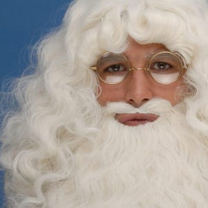 Könnte beinhalten: Nahaufnahme einer Person in einem Weihnachtsmannkostüm mit weißer Perücke, Bart und Schnurrbart. Die Person trägt auch eine runde, goldgerahmte Brille. Der Hintergrund ist einfarbig blau.
