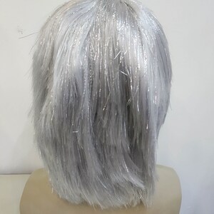 May include: A gray wig with silver tinsel strands, styled in a short, layered cut. The wig is displayed on a mannequin head. The hair has a slightly textured appearance, with the silver strands adding a touch of sparkle.