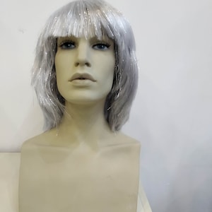 May include: A mannequin head with a silver wig. The wig has short, layered strands with silver tinsel accents. The mannequin has a neutral skin tone, blue eyes, and a neutral expression. The background is a plain white wall.