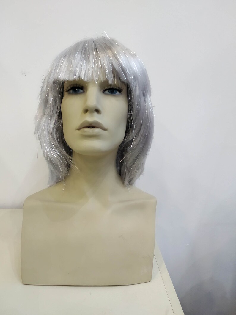 May include: A mannequin head with a silver wig. The wig has short, layered strands with silver tinsel accents. The mannequin has a neutral skin tone, blue eyes, and a neutral expression. The background is a plain white wall.