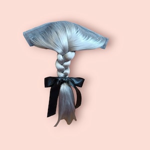 Könnte beinhalten: Eine silbergraue geflochtene Haarverlängerung mit einer schwarzen Satin-Schleife. Die Haarverlängerung ist an einem grauen Netzclip befestigt.