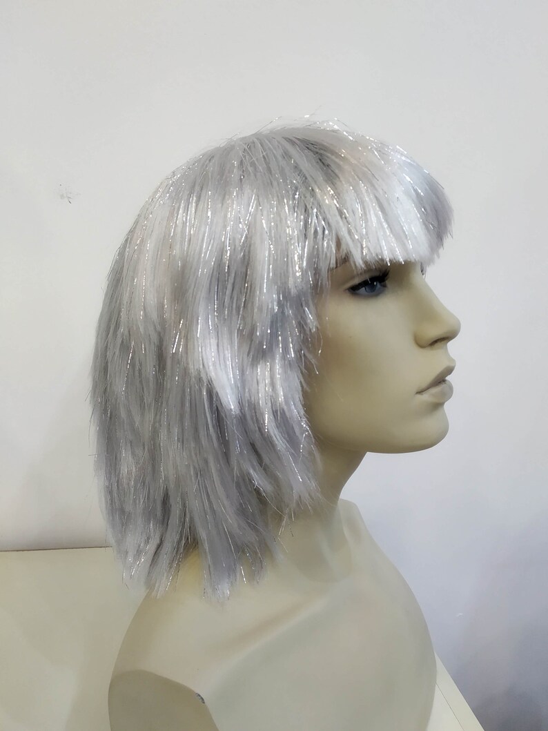 May include: A silver wig with a blunt fringe, displayed on a mannequin head. The wig has a short, layered style with strands of silver tinsel throughout, creating a shimmering effect. The mannequin has a neutral skin tone and is set against a white background.