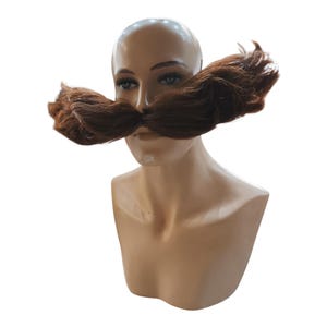 May include: A brown, bushy mustache prop is displayed on a mannequin head. The mustache is thick and covers the upper lip, with a slightly curved shape. The mannequin has a neutral skin tone and is set against a white background.