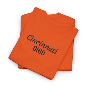 Puede incluir: Dos camisetas naranjas apiladas. La camiseta superior tiene las palabras "Cincinnati OHIO" impresas en negro. Las camisetas están dobladas y sobre un fondo blanco. Las camisetas son de color naranja brillante.