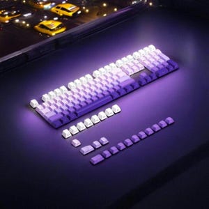 May include: A purple and white mechanical keyboard with a gradient of purple keys. The keyboard is on a dark surface and includes a set of extra keys.