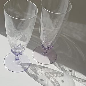 May include: Two clear glass flutes featuring etched palm leaf patterns and circular accents. Each glass has a light purple tinted stem and a circular base. The glasses are positioned upright, casting distinct shadows on the surface below.