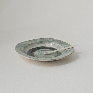 May include: A ceramic plate with an organic shape and a mottled green and grey glaze. A small, white spoon is placed on the plate. The plate has a darker, circular design in the centre. This item is suitable for serving small portions. The plate's diameter is approximately 20 cm.