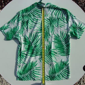 May include: White button-up shirt featuring a bold green palm leaf design. A yellow tape measure indicates a length of approximately 74 cm. The shirt is suspended from a green hanger.