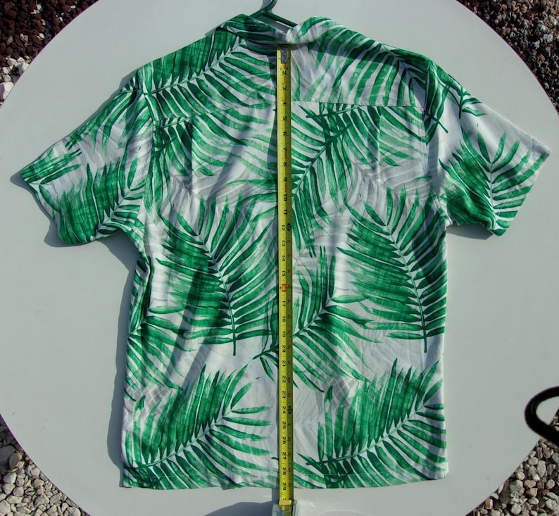 May include: White button-up shirt featuring a bold green palm leaf design. A yellow tape measure indicates a length of approximately 74 cm. The shirt is suspended from a green hanger.