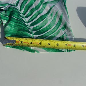 May include: Close-up of a section of fabric with a green and white tropical leaf pattern. A yellow tape measure shows the fabric is approximately 25 cm wide.