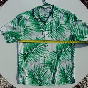 May include: Short-sleeved button-down shirt with a white background and a vibrant green palm leaf pattern. The shirt is measured with a tape measure, showing its size. The shirt is hanging on a green hanger.