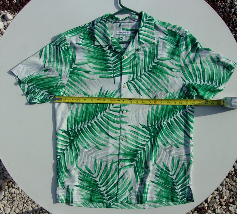 May include: Short-sleeved button-down shirt with a white background and a vibrant green palm leaf pattern. The shirt is measured with a tape measure, showing its size. The shirt is hanging on a green hanger.