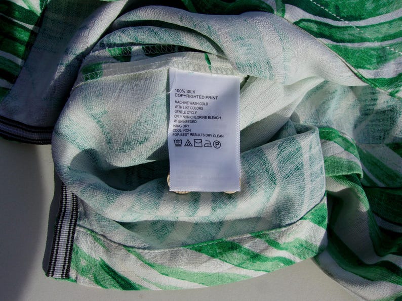May include: Close-up of a silk fabric with a green and white striped pattern. A white tag with care instructions is attached. The tag reads "100% Silk Copyrighted Print". The fabric is soft and appears to be part of a garment.