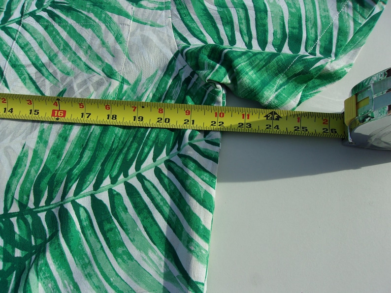 May include: A white and green tropical print shirt with a palm leaf design. A yellow measuring tape is placed across the fabric, showing a measurement of approximately 66 cm.