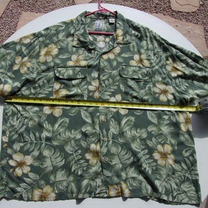 May include: Green button-down shirt with a floral pattern. The shirt features a tropical design with large white and yellow flowers and green leaves. The shirt is approximately 30 inches wide.