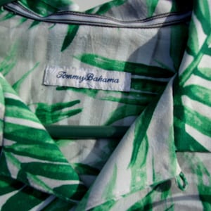 May include: Close-up of a white and green patterned shirt with a tropical leaf design. The shirt has a collar and a label that reads "Tommy Bahama". The shirt is hanging on a green hanger.
