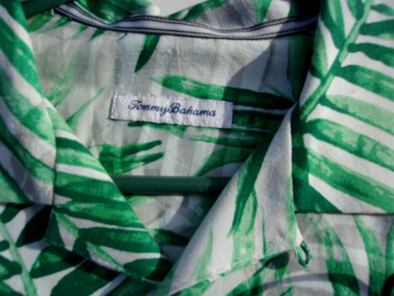 May include: Close-up of a white and green patterned shirt with a tropical leaf design. The shirt has a collar and a label that reads "Tommy Bahama". The shirt is hanging on a green hanger.