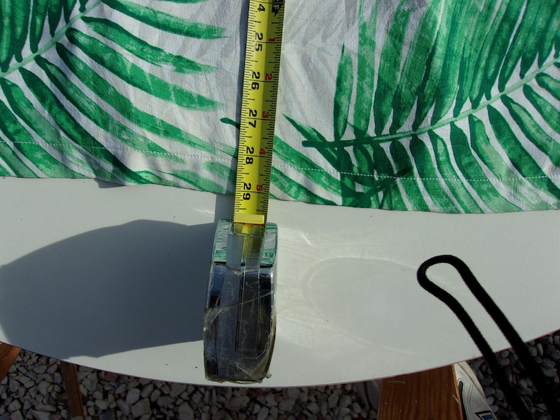May include: White fabric with a green palm leaf pattern is measured with a yellow tape measure. The tape measure shows a measurement of 61 to 74 cm. The fabric is on a white surface.