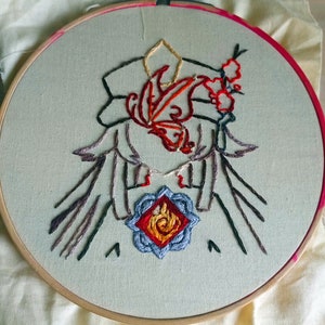 May include: Embroidered hoop art featuring a stylised figure with a red and orange butterfly design, holding a blue and red flower with a flame inside. The figure is surrounded by black and brown lines.