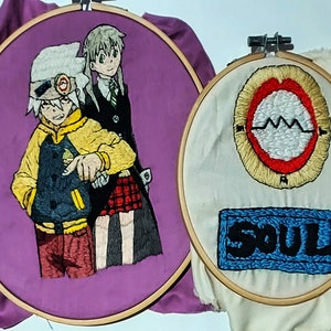 May include: Two embroidered hoop art pieces featuring characters from the anime Soul Eater. The first hoop features Soul Eater and Maka Albarn, with Soul Eater wearing a yellow jacket and Maka wearing a black and red dress. The second hoop features a red and white symbol with the word "SOUL" embroidered in blue.