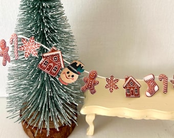 May include: A festive Christmas scene with a small green tree and a garland of wooden decorations. The garland includes gingerbread men, houses, snowflakes, and candy canes. The scene is set on a small, cream-coloured table.
