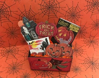 May include: A miniature cardboard box filled with Halloween decorations, including a ghost, a pumpkin, and a sign that says "Trick or Treat". The box is sitting on a red and black background with a spiderweb pattern.