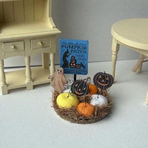 May include: A miniature Halloween display featuring a blue sign that reads The Black Hat PUMPKIN PATCH Locally Grown WITCHING PUMPKINS SINCE 1692. The display includes a small ghost cutout, several fabric pumpkins in yellow, orange, and white, and two black jack-o'-lantern cutouts nestled in a bed of brown dried grass.