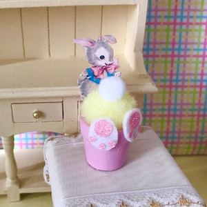 May include: A miniature Easter-themed decoration featuring a pink pot with a yellow, fluffy center. A paper cutout of a gray bunny with a pink bow is attached. White and pink glitter bunny feet are on the pot's side. The decoration is on a white surface.