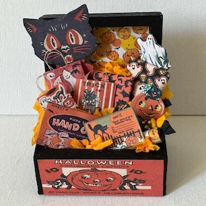 Puede incluir: Una pequeña caja negra llena de decoraciones de Halloween. Incluye un recorte de gato negro, bolsas de caramelos, un botón de calabaza y recortes de cartón vintage. Papel de seda naranja y amarillo añade color.