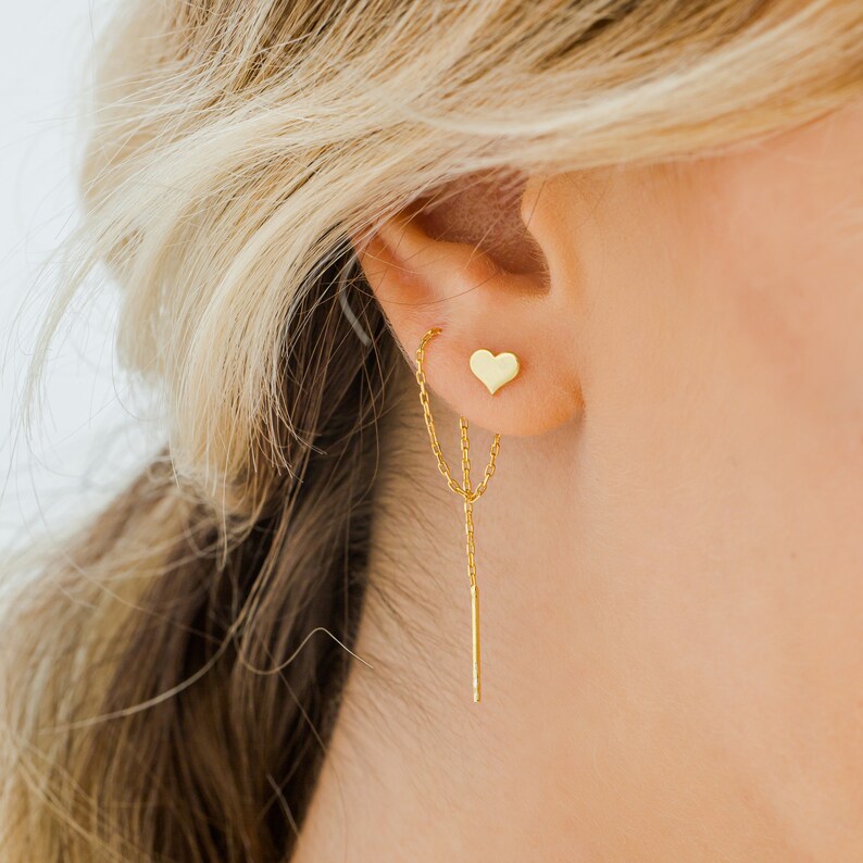 May include: Gold heart shaped earring with a delicate chain dangling from the bottom. The chain has a small gold bar at the end.
