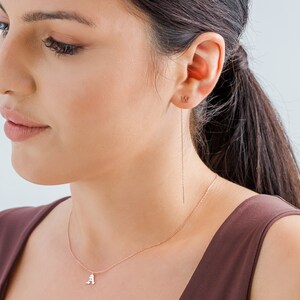 May include: A rose gold necklace with a delicate chain and a small letter 'A' pendant. The model is also wearing a rose gold chain earring with a small pendant.