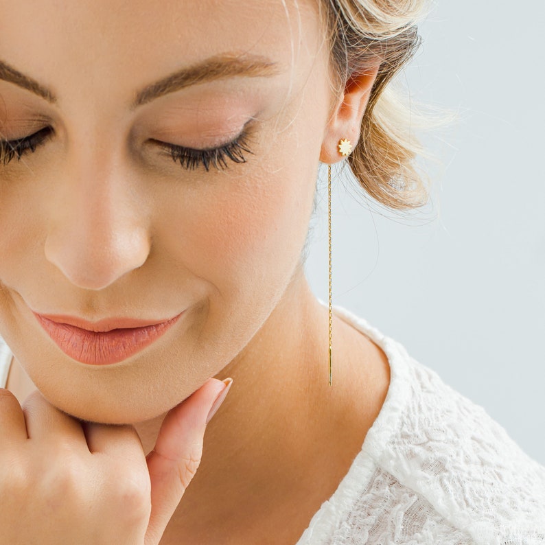 May include: A woman wearing a white top with a gold chain earring featuring a small star charm.