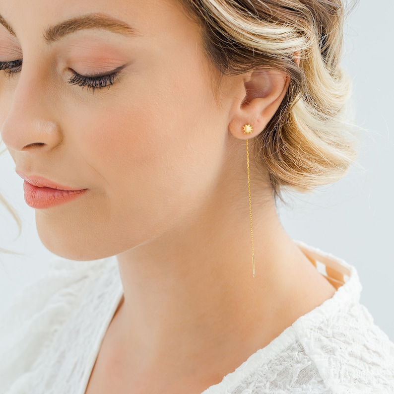 May include: Gold star-shaped earrings with a long, thin chain dangling from the bottom. The earrings are worn by a person with blonde hair.