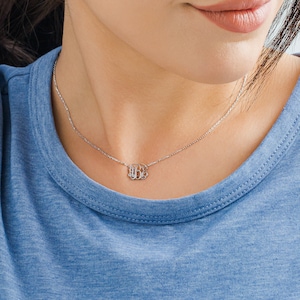 Puede incluir: Un collar de cadena de plata con un pequeño y delicado colgante de monograma. El colgante presenta tres letras entrelazadas en una fuente de escritura clásica.