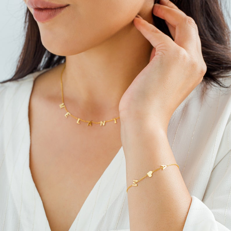 May include: A gold necklace with the name "MELANI" spelled out in individual letters. A matching gold bracelet with the name "MIA" spelled out in individual letters and a small heart charm.