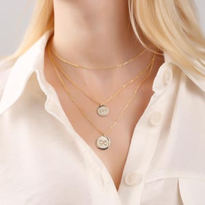 May include: Three gold necklaces of different lengths. The top necklace is a simple chain. The second necklace has a small, round pendant with the letters "BVO". The third necklace has a larger, round pendant with an infinity symbol.