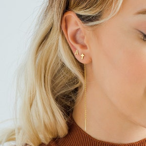 May include: Gold earrings with a small cross and the letter 'A' on one ear and a long chain earring with a thin gold bar on the other ear.